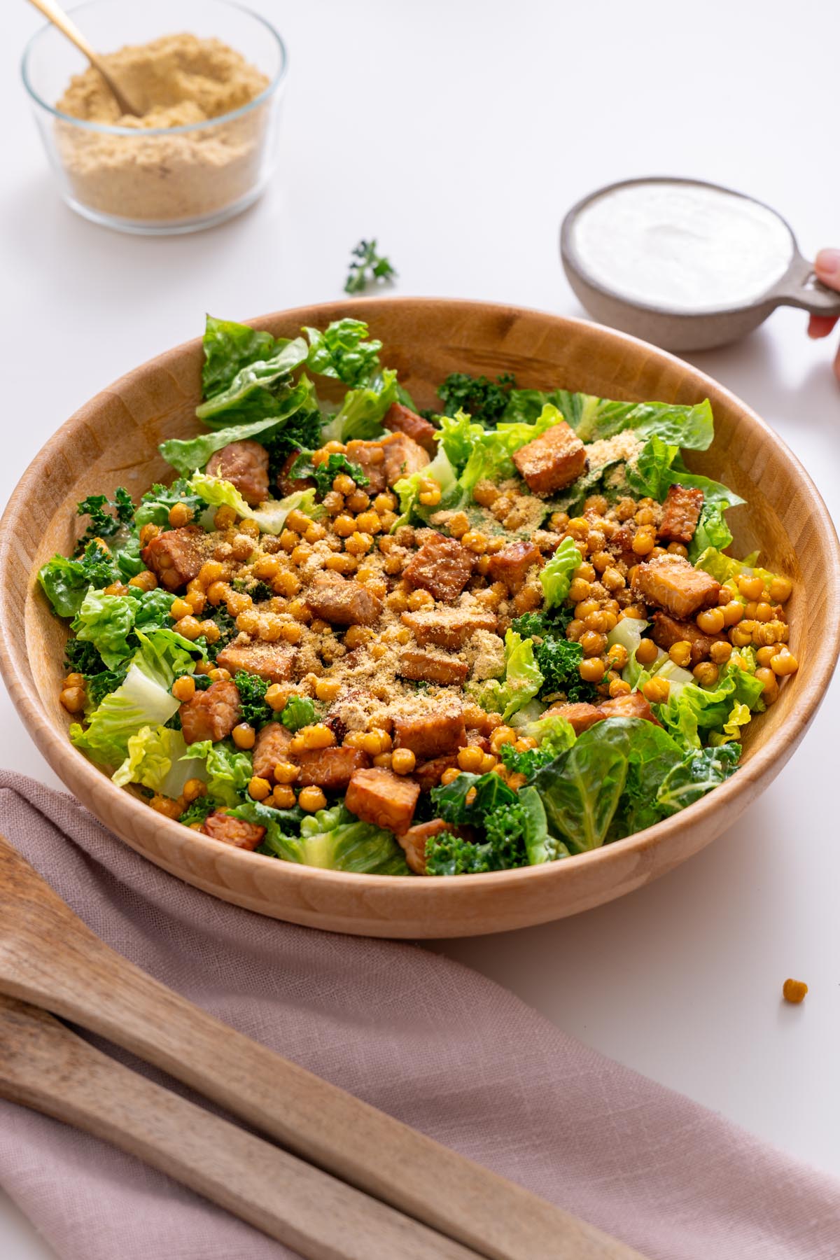 Large wooden bowl of vegan Caesar salad with romaine, kale, crispy tempeh, chickpeas, and parmesan, with dressing being held nearby.