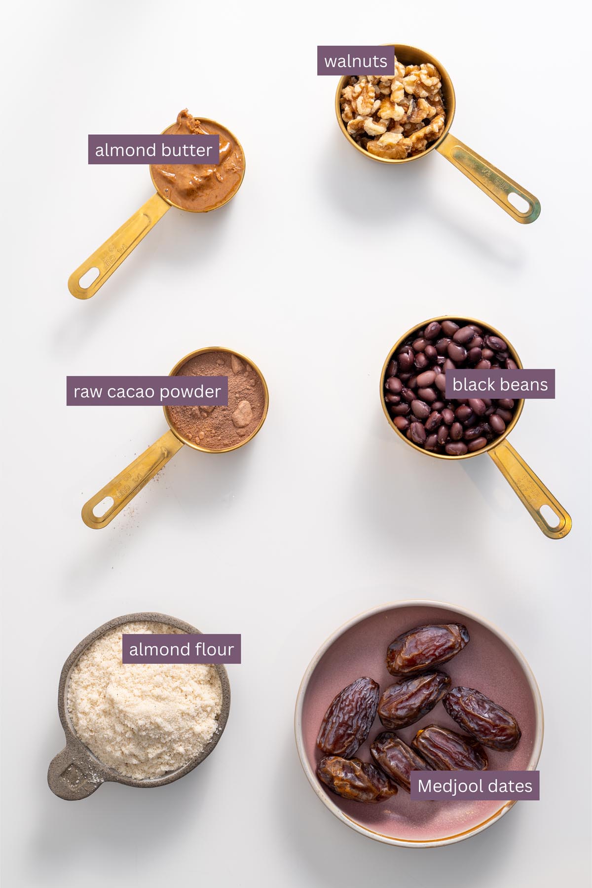 Vibrant ingredients for fudgy no-bake brownies on a white surface, including almond butter, walnuts, black beans, cacao powder, almond flour, and Medjool dates