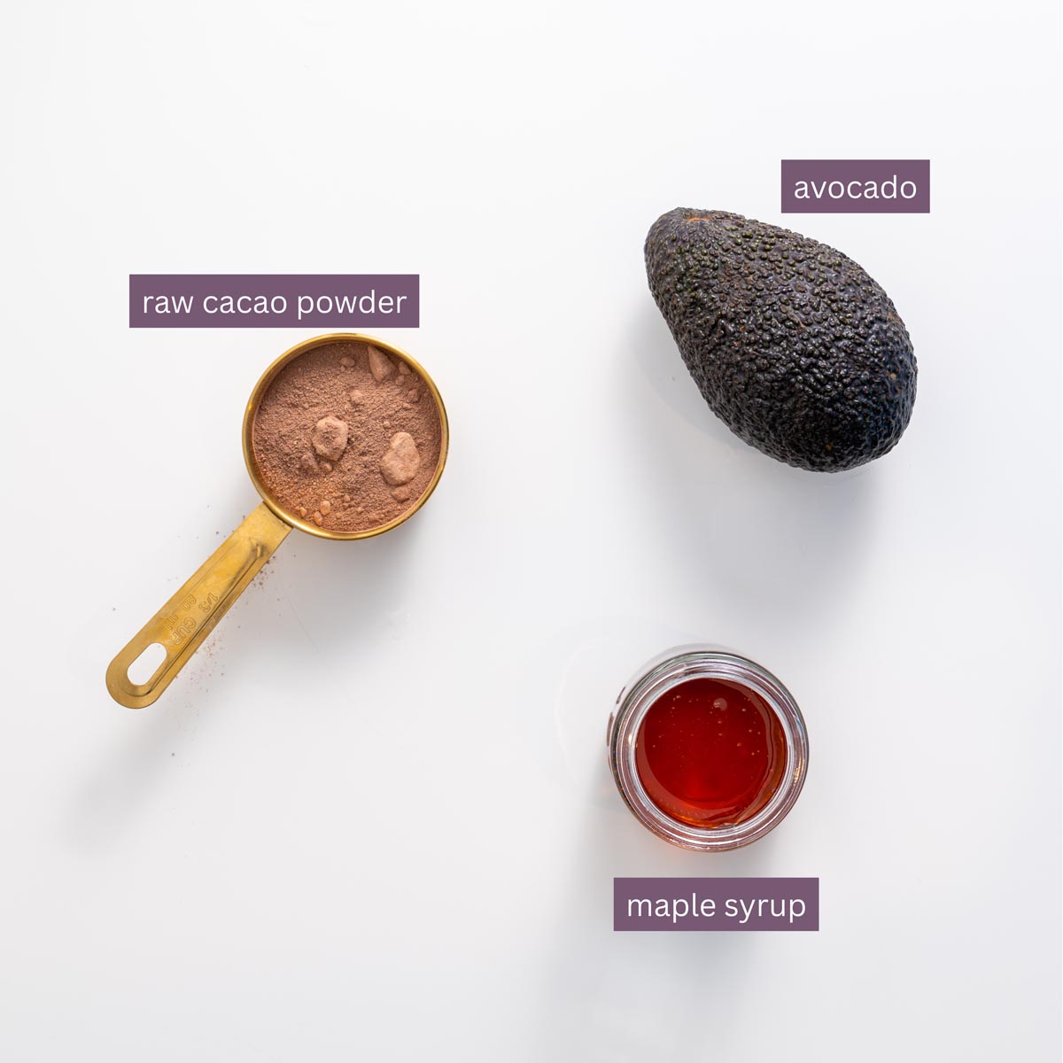 Vibrant ingredients for avocado ganache on a white surface, including avocado, cacao powder, and maple syrup