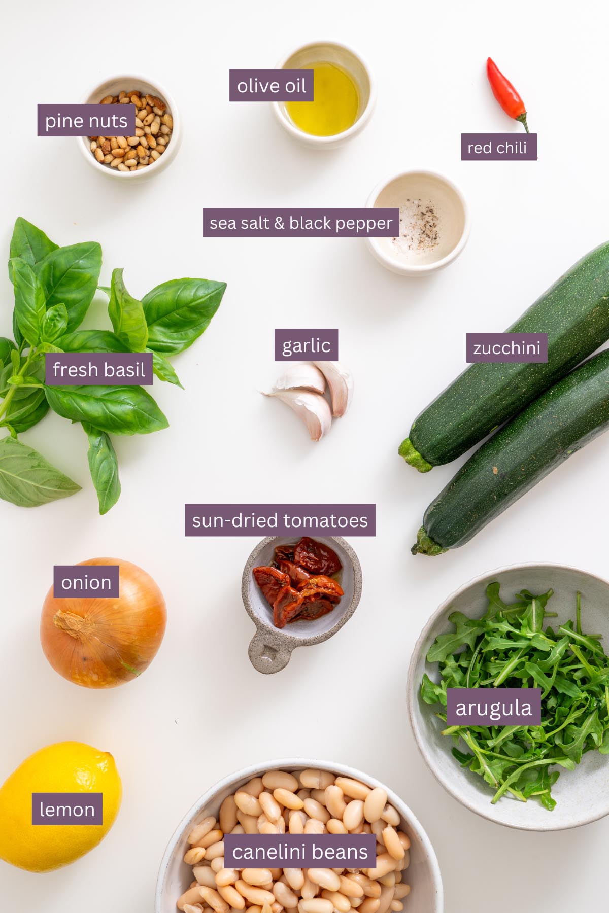 Các thành phần chính cho công thức đậu zucchini bao gồm zucchini, đậu cannellini, húng quế, chanh, tỏi, hành tây, ớt, cà chua khô, arugula, hạt thông, dầu ô liu và gia vị trên bề mặt trắng