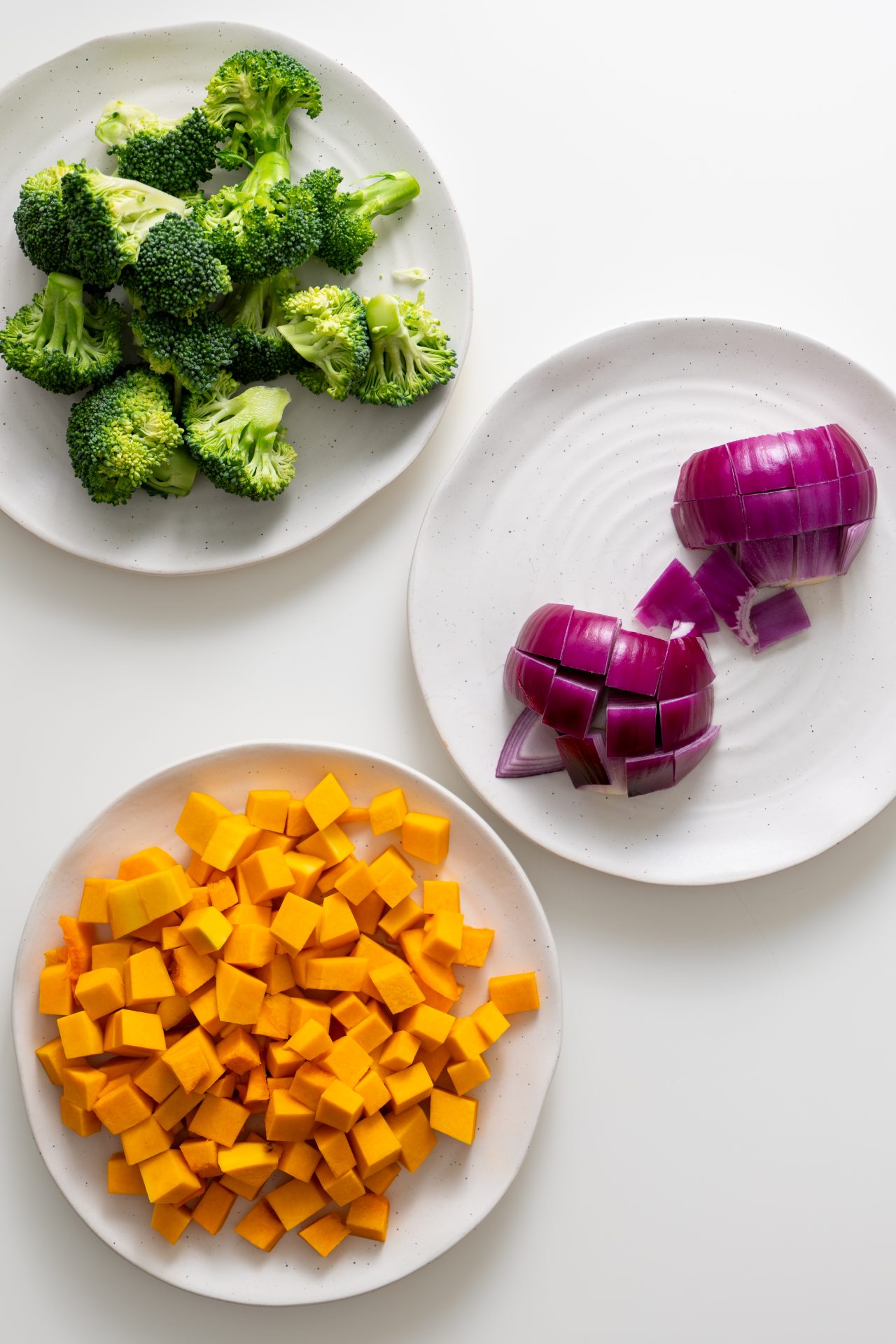Fresh broccoli florets, cubed pumpkin, and chopped red onion on white plates, ready for roasting