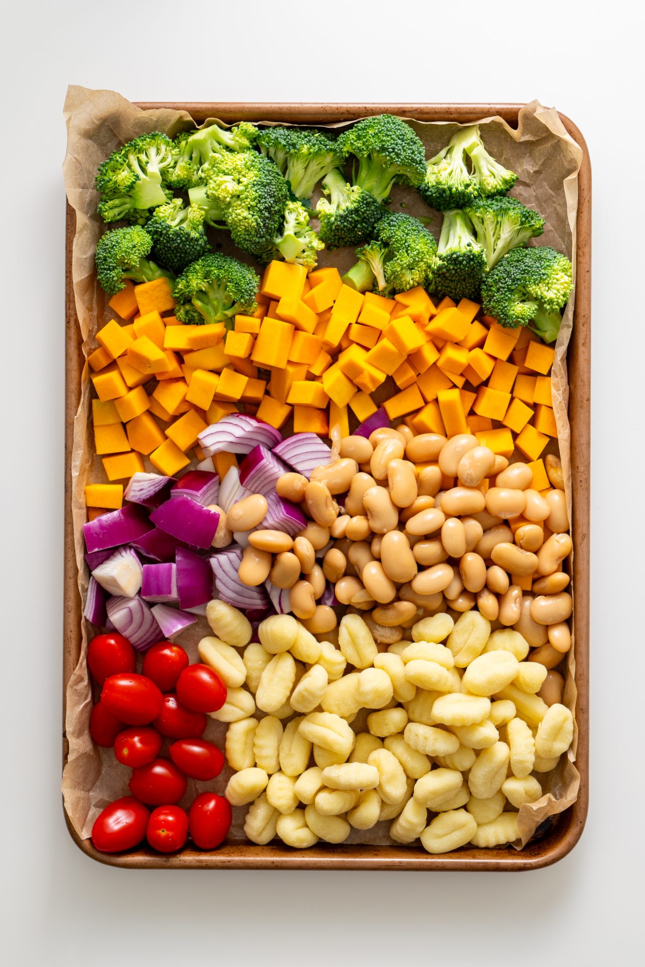Raw broccoli, pumpkin cubes, cherry tomatoes, red onion, butter beans, and gnocchi arranged on a parchment-lined sheet pan