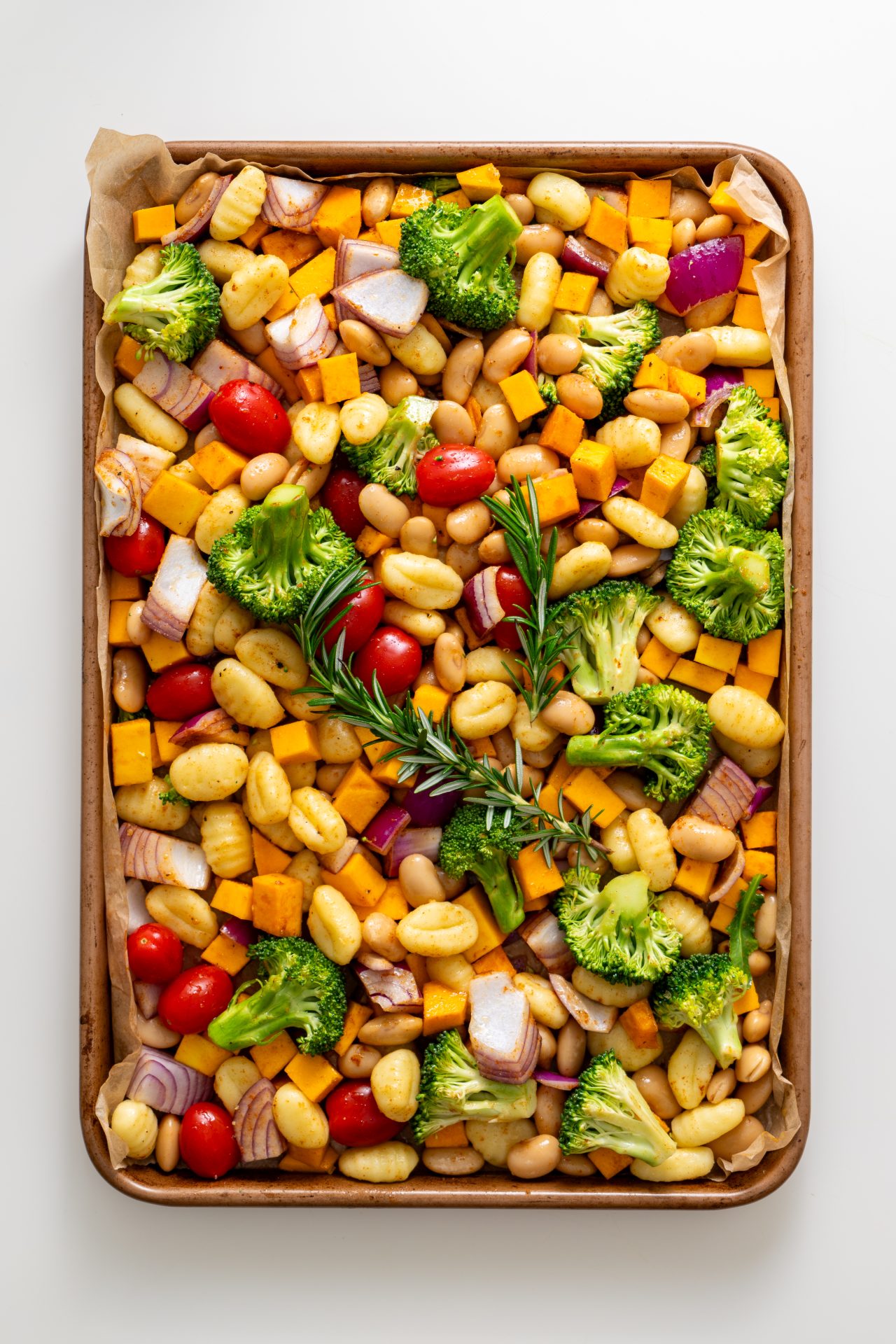 Seasoned gnocchi, pumpkin, broccoli, onion, tomatoes, butter beans, and rosemary sprigs spread on a parchment-lined baking sheet