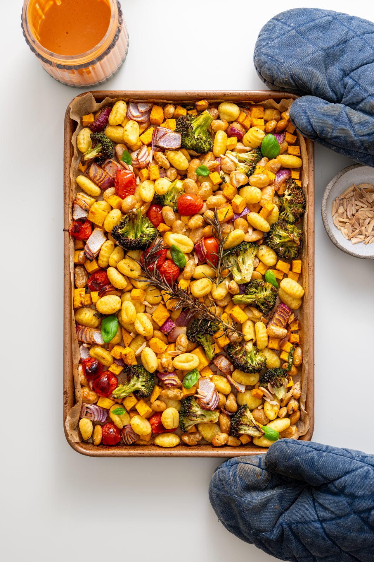 Freshly baked sheet pan gnocchi with roasted vegetables and rosemary sprigs, held with oven mitts