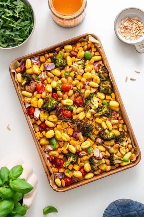 Overhead view of roasted gnocchi with vegetables on a baking tray, surrounded by arugula, tahini sauce, and almonds