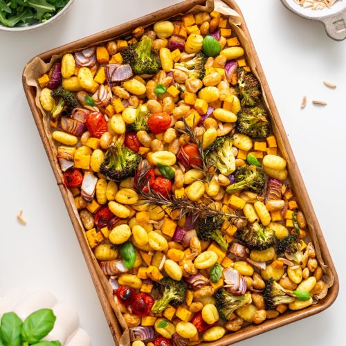 Overhead view of roasted gnocchi with vegetables on a baking tray, surrounded by arugula, tahini sauce, and almonds