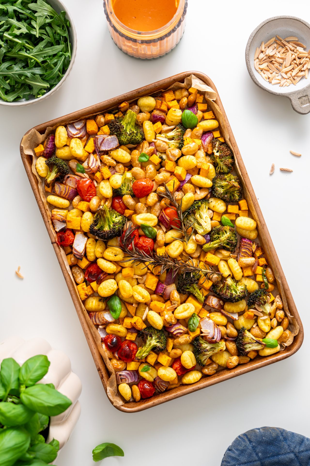 Overhead view of roasted gnocchi with vegetables on a baking tray, surrounded by arugula, tahini sauce, and almonds