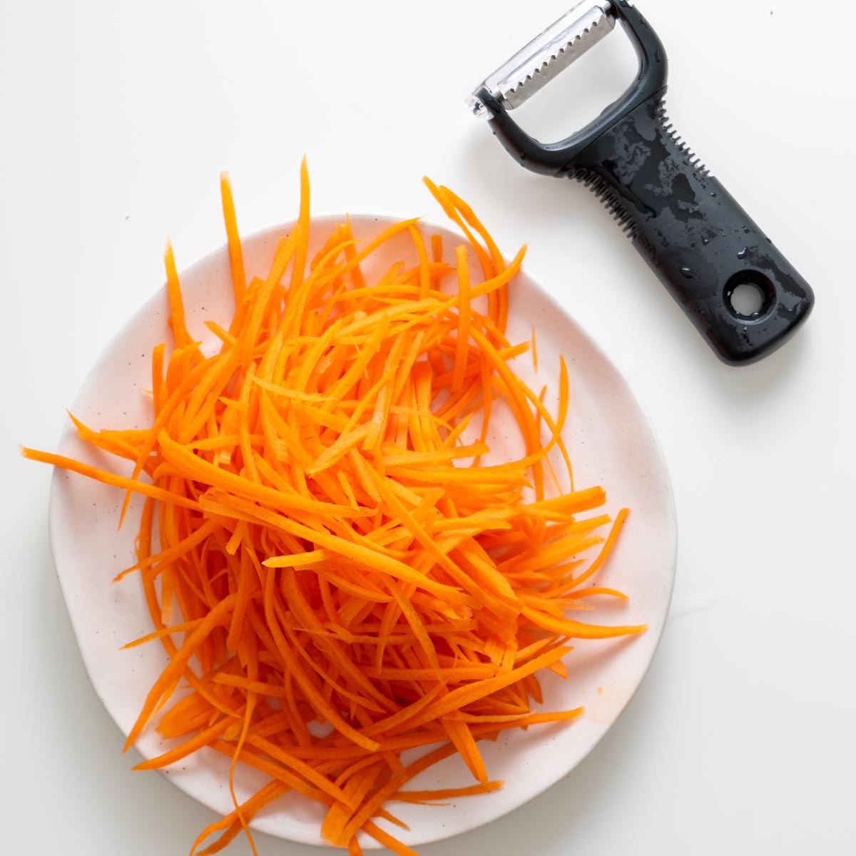 Julienned carrots on a plate with a julienne peeler for peanut vermicelli salad jars