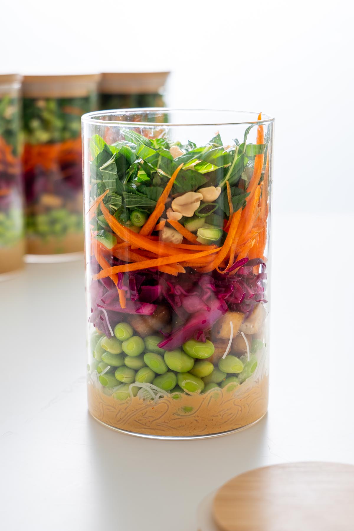 Close-up of a layered peanut vermicelli salad jar with noodles, tofu, edamame, cabbage, carrots, mint, scallions, and peanuts