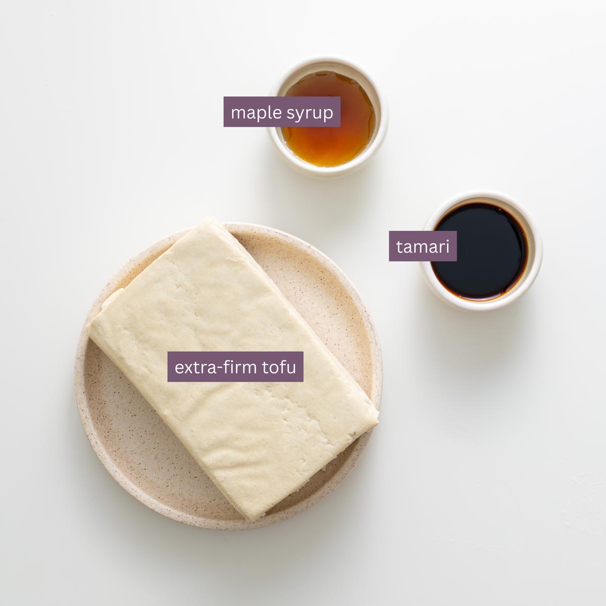 Block of extra-firm tofu with small bowls of tamari and maple syrup on a white surface