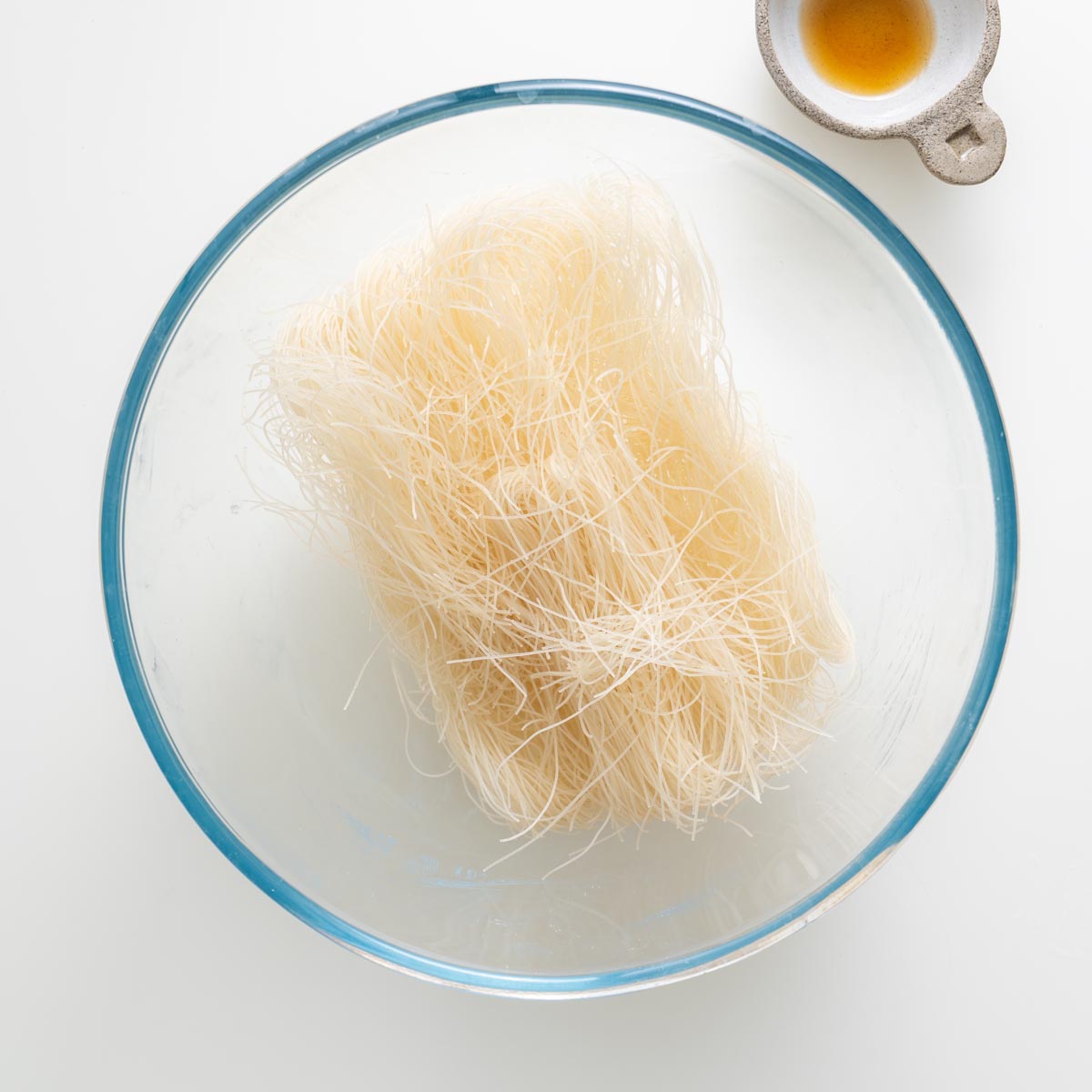 Dry rice vermicelli noodles in a glass bowl with a small dish of sesame oil beside it