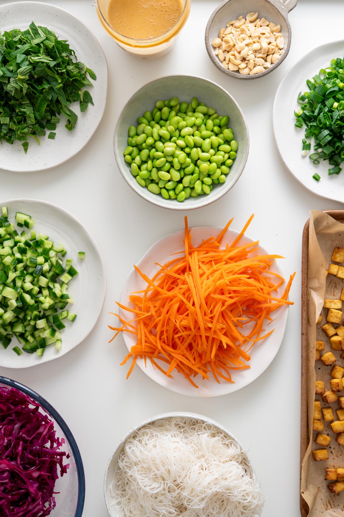 Prepared salad components including chopped mint, edamame, cucumbers, shredded carrots, cabbage, scallions, peanuts, baked tofu, noodles, and peanut dressing arranged on plates and bowls