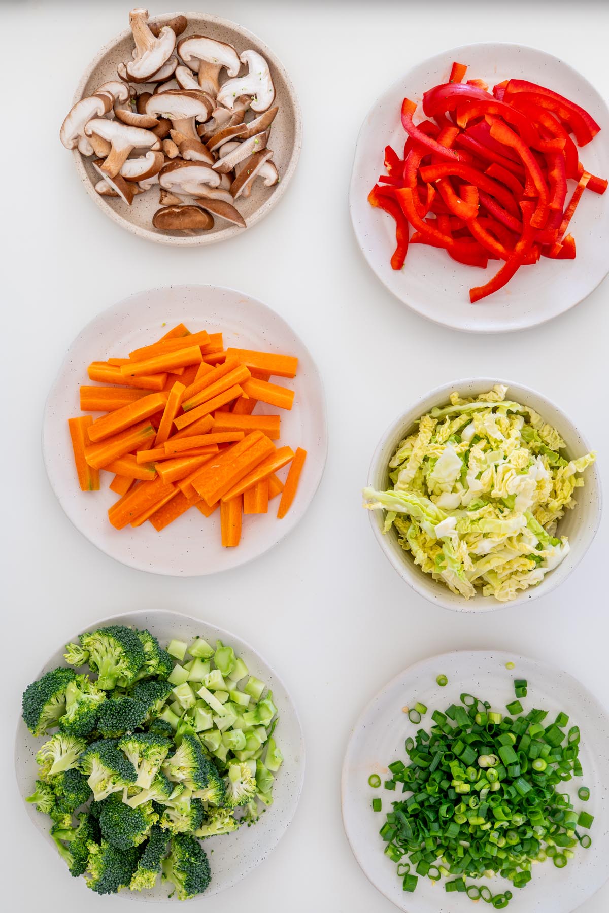 Plates of prepared vegetables including mushrooms, red bell pepper, carrots, cabbage, broccoli, and scallions on a white surface