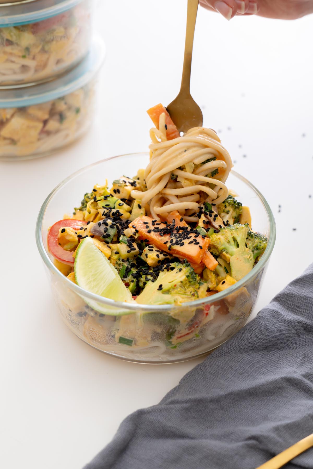 Glass meal prep bowl of veggie noodle stir fry with tofu, vegetables, black sesame seeds, and a fork twirling noodles