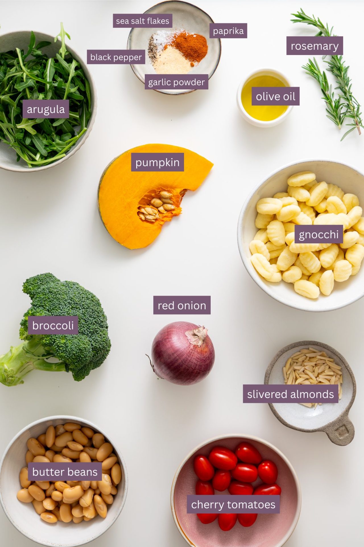 Vibrant ingredients for sheet pan gnocchi with roasted veggies on a white surface, including pumpkin, broccoli, cherry tomatoes, butter beans, gnocchi, red onion, herbs, spices, and almonds