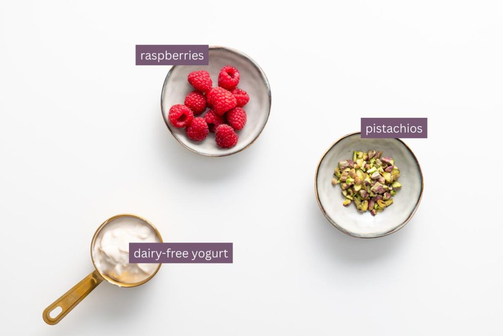 Vibrant ingredients for a blended matcha chia pudding including raspberries, pistachios, and dairy-free yogurt on a white surface.