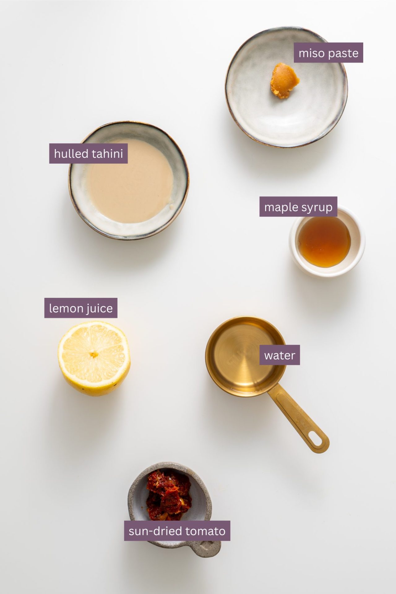 Vibrant dressing ingredients for tahini sauce on a white surface, including tahini, lemon, maple syrup, miso paste, sun-dried tomato, and water
