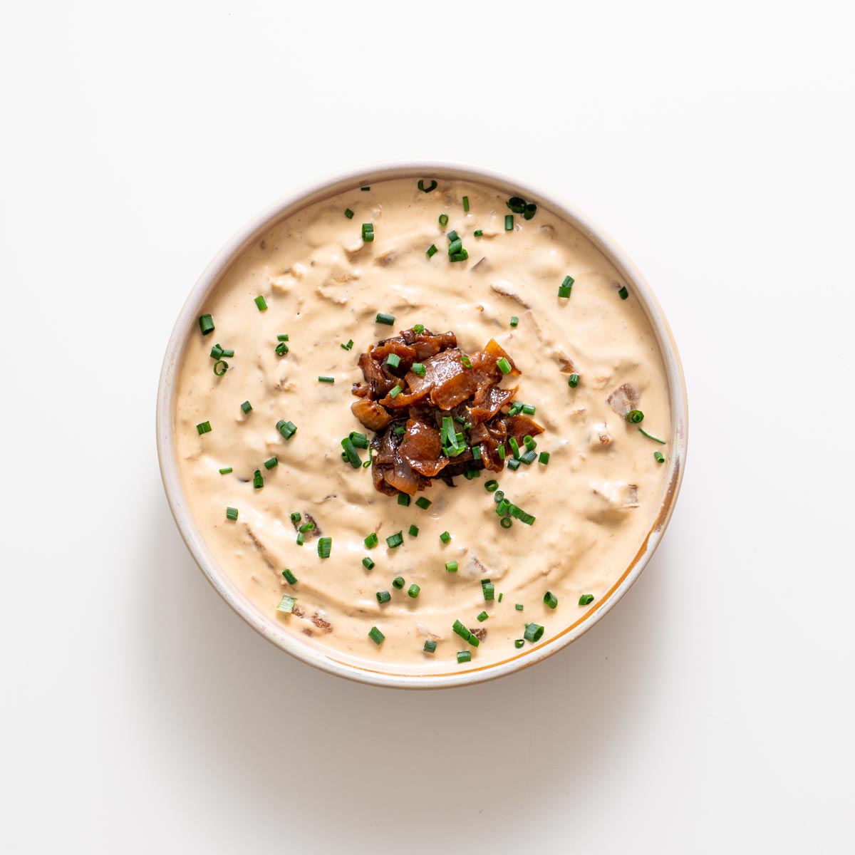 French onion dip topped with caramelized onions and chopped chives on a white surface.