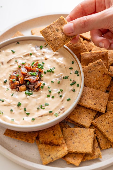 Sarah Cobacho holding an almond flour rosemary cracker dipped into creamy French onion dip, surrounded by crackers on a plate.