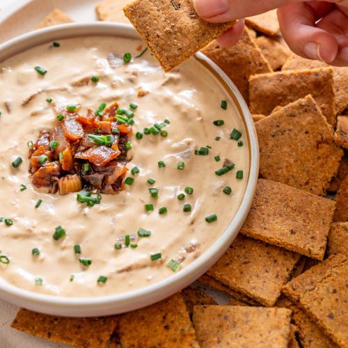 Sarah Cobacho holding an almond flour rosemary cracker dipped into creamy French onion dip, surrounded by crackers on a plate.