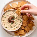 Sarah Cobacho holding a baked almond flour rosemary cracker above creamy onion dip, surrounded by crackers on a white plate.
