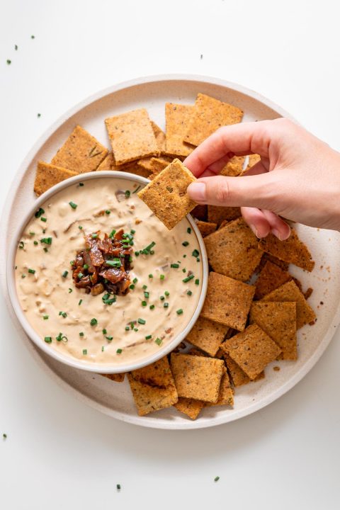 Sarah Cobacho holding a baked almond flour rosemary cracker above creamy onion dip, surrounded by crackers on a white plate.