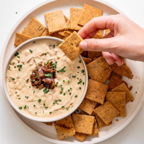 Sarah Cobacho holding a baked almond flour rosemary cracker above creamy onion dip, surrounded by crackers on a white plate.