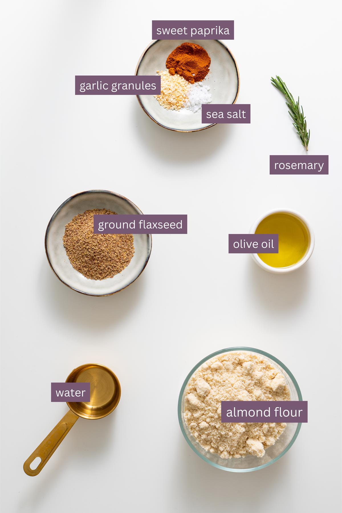 Vibrant labeled ingredients for almond flour rosemary crackers on a white surface, including almond flour, ground flaxseed, garlic granules, sweet paprika, sea salt, rosemary, olive oil, and water.