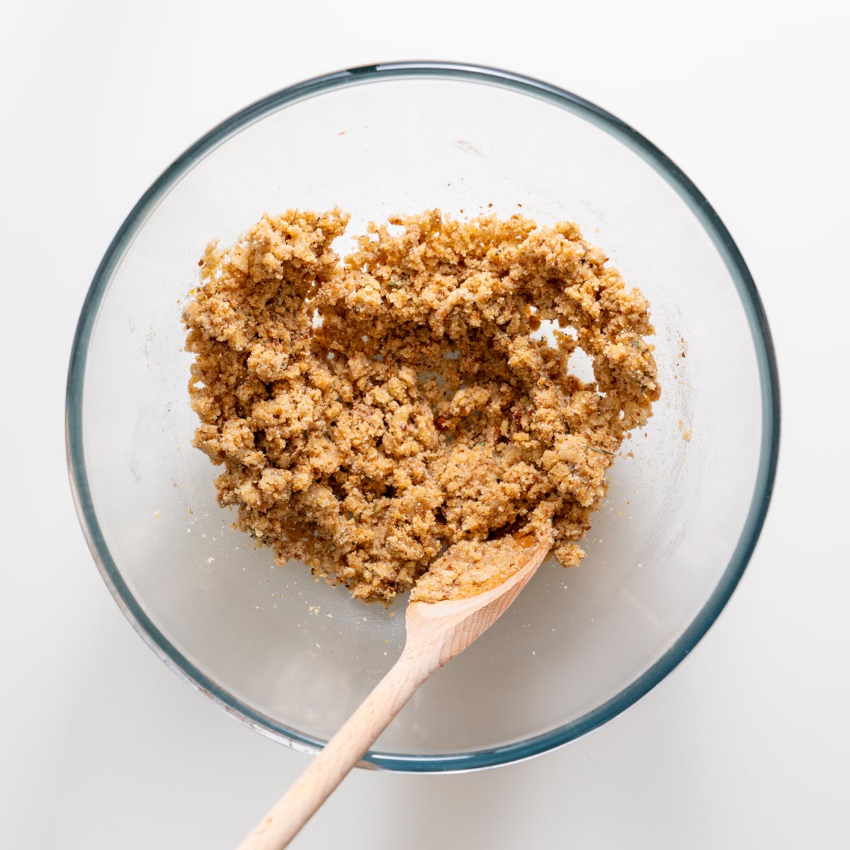 Crumbly almond flour rosemary cracker dough being mixed with a wooden spoon in a glass bowl.