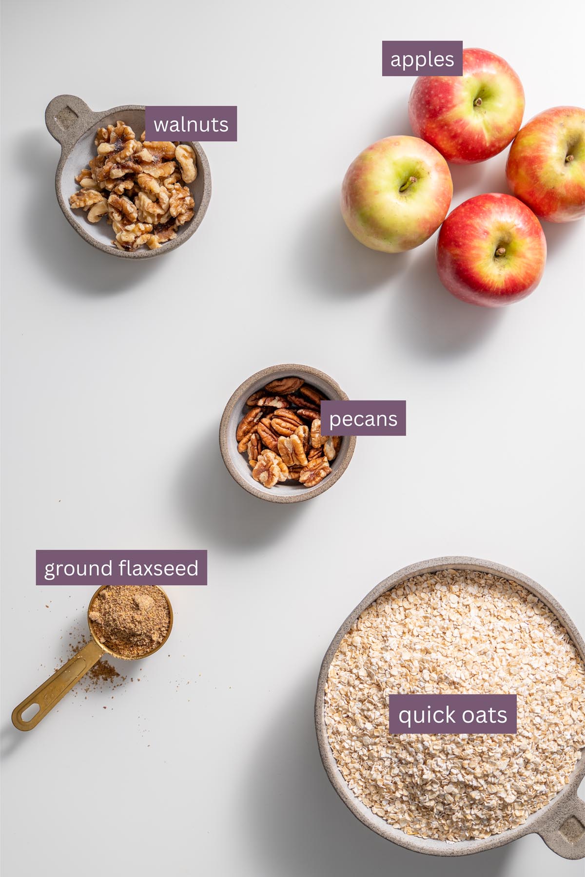 Labeled ingredients for apple pie baked oats including apples, walnuts, pecans, ground flaxseed and quick oats on a white surface.