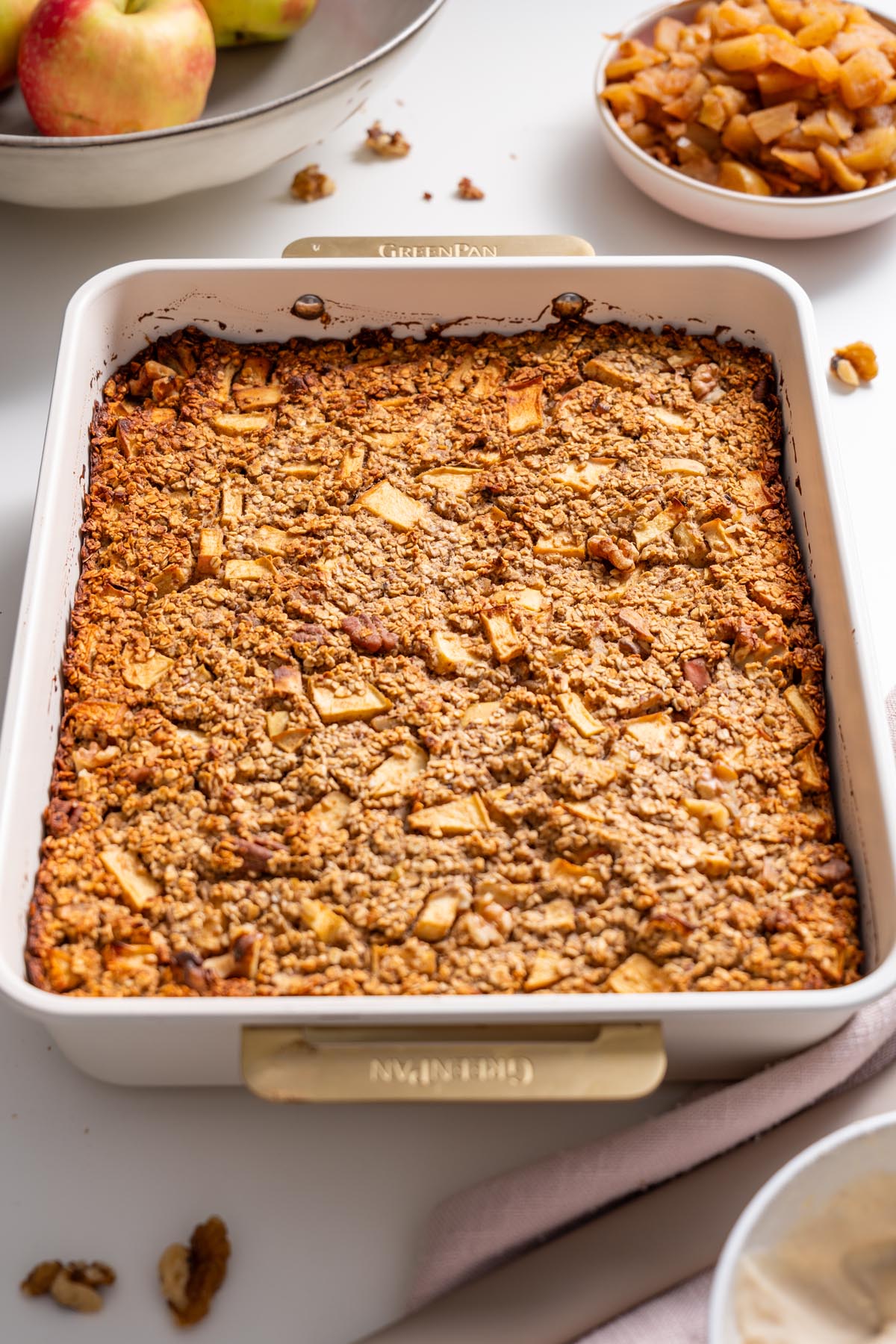 Baking dish of apple pie baked oats with diced apples and a bowl of stewed apples nearby.