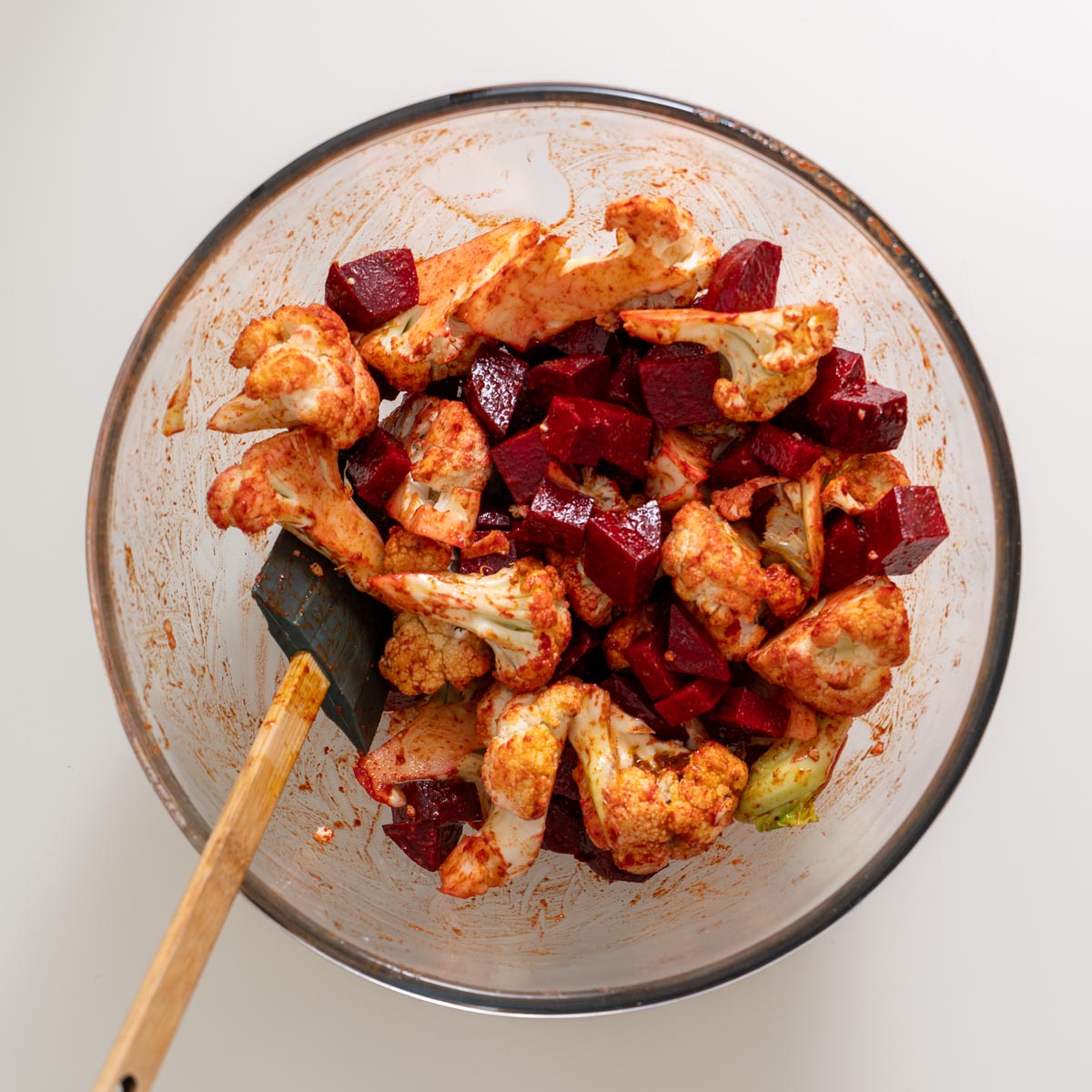 Cauliflower and beetroot tossed in harissa marinade in a large glass mixing bowl with a spatula