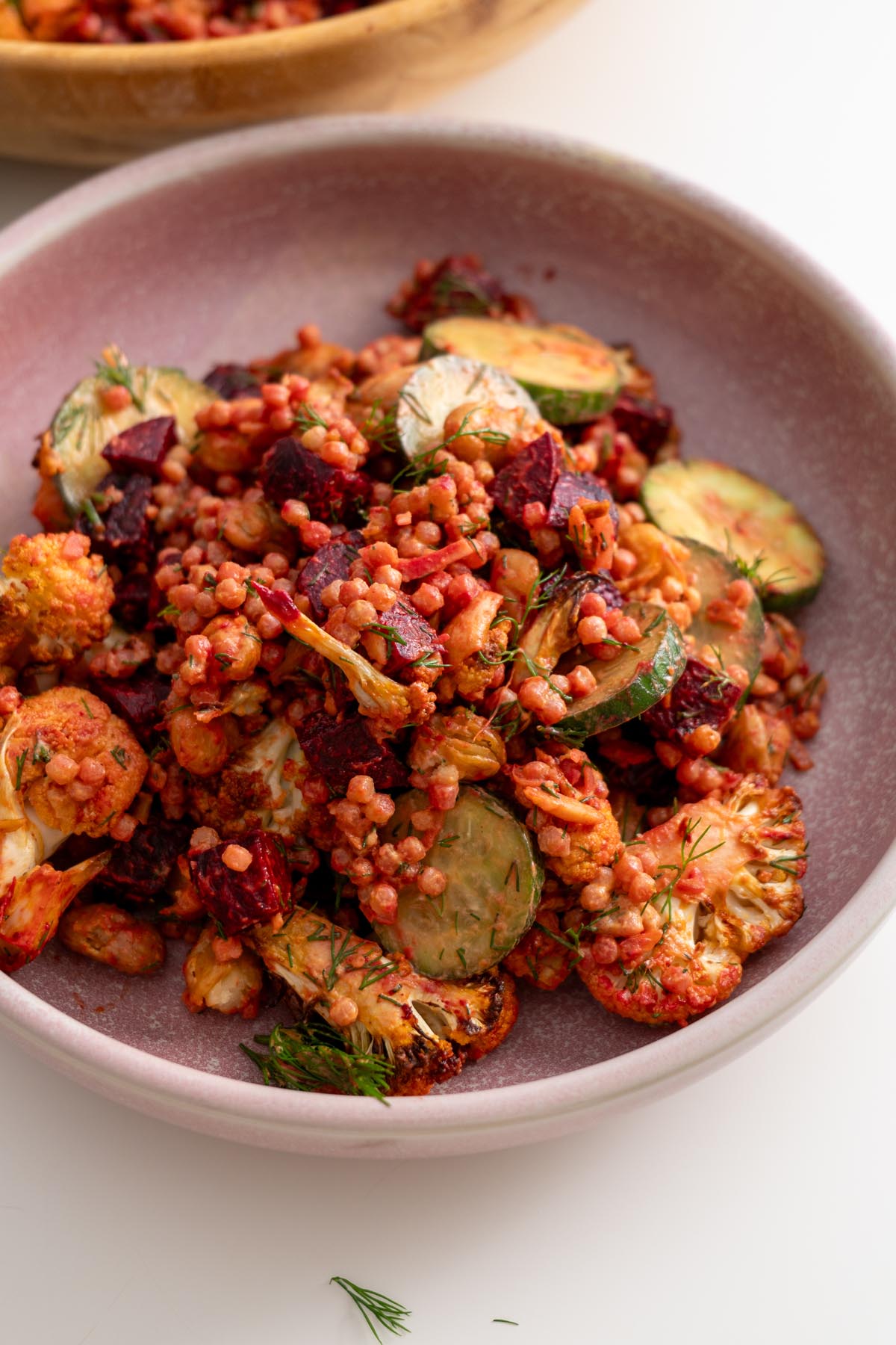 Single serving of harissa cauliflower beetroot salad in a pink bowl with roasted vegetables, cucumber, and couscous