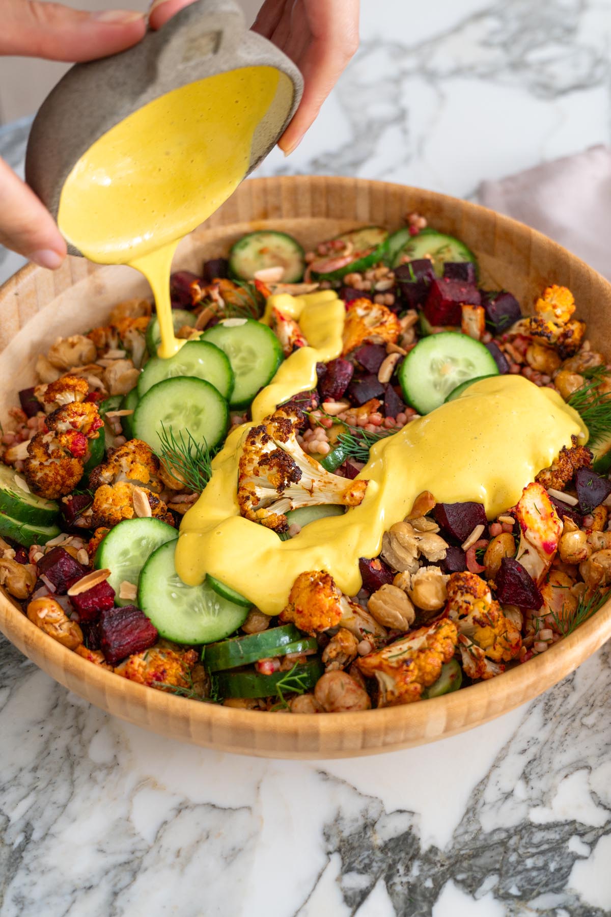 Sarah Cobacho pouring golden tahini turmeric dressing over a harissa cauliflower beetroot salad in a wooden bowl