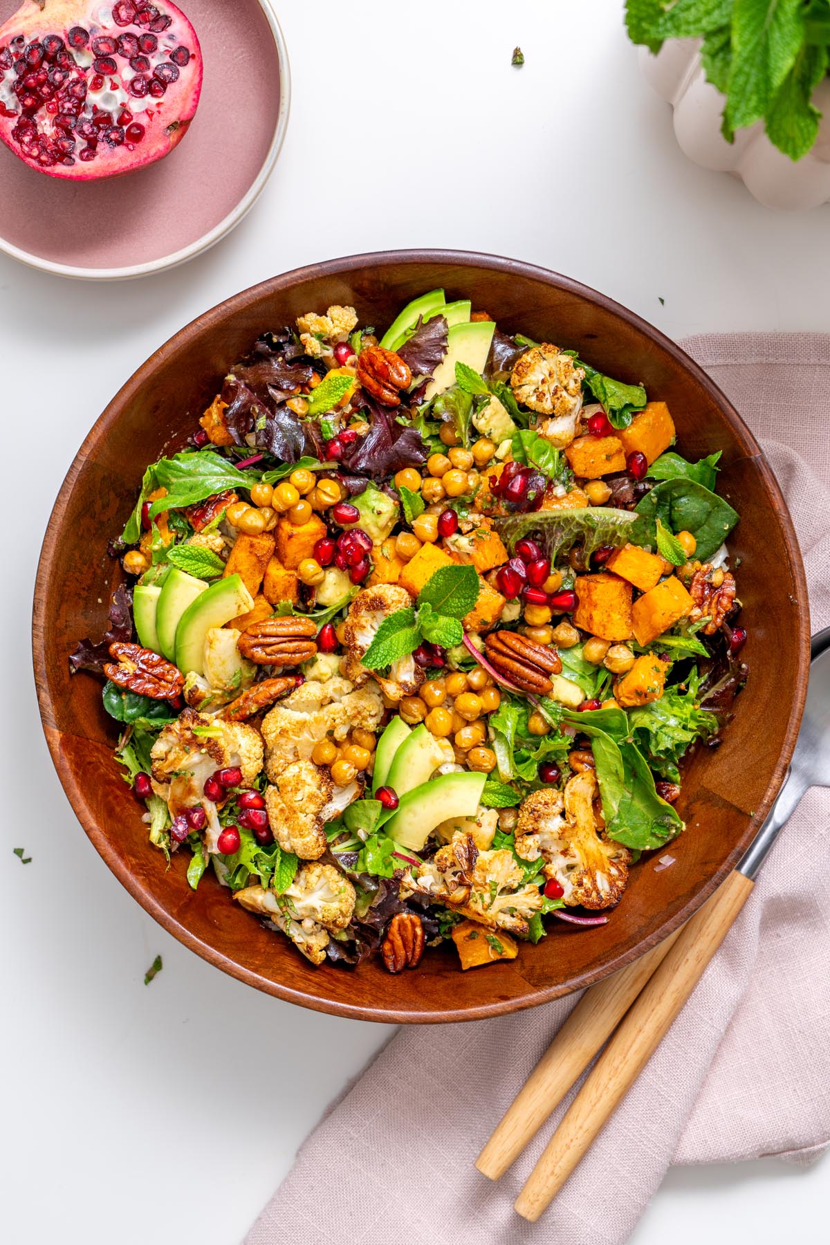 Overhead view of festive maple salad with roasted vegetables, chickpeas, avocado, pomegranate seeds, candied pecans and leafy greens in a wooden bowl