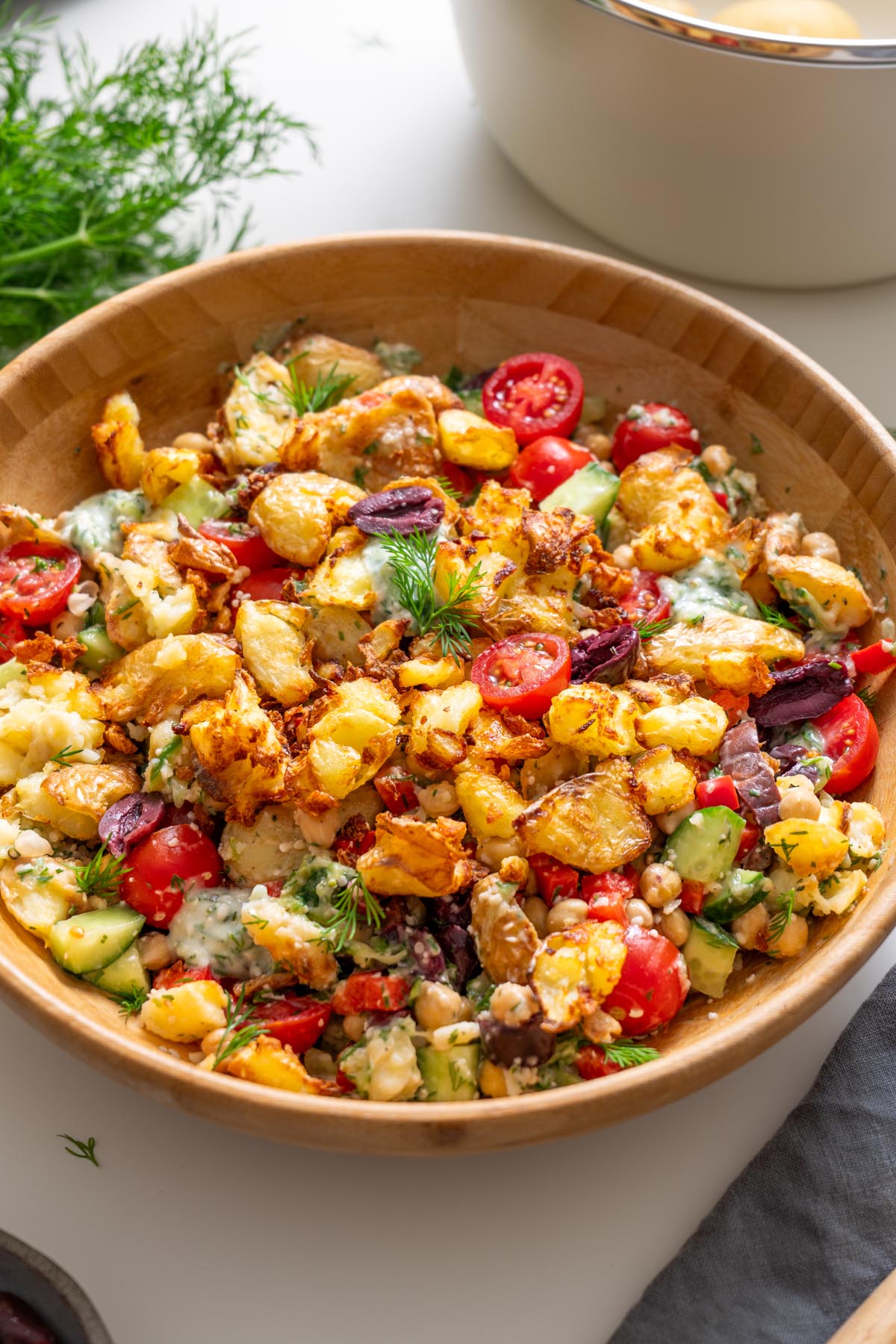 Greek-style crispy potato salad with tzatziki in a wooden bowl surrounded by dill and kitchenware.