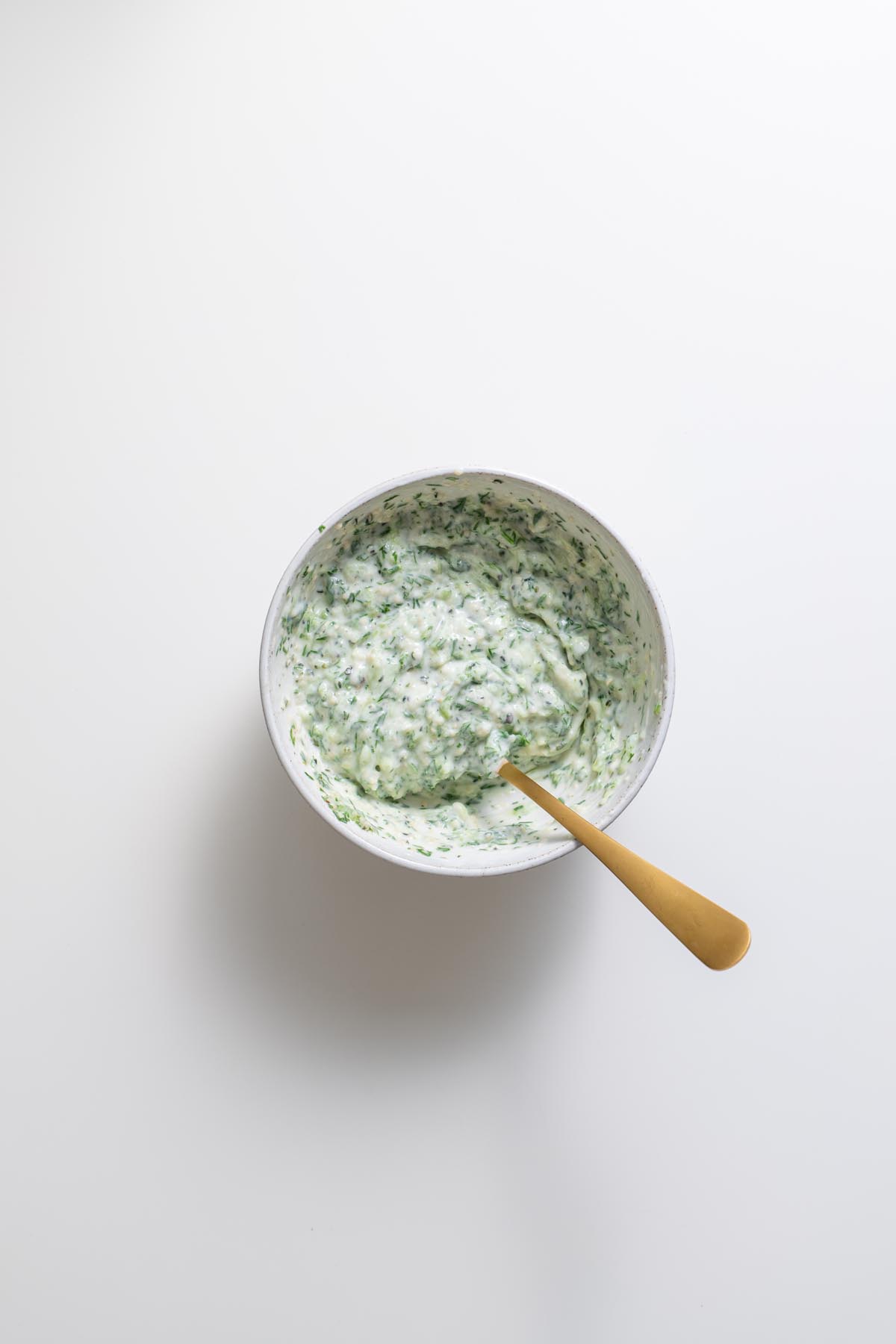 Creamy dairy-free tzatziki sauce in a white bowl with a gold spoon on a white background.