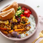 Bowl of Mediterranean sheet pan pita bowls with tzatziki, roasted veggies, olives, and pita on a light pink napkin.