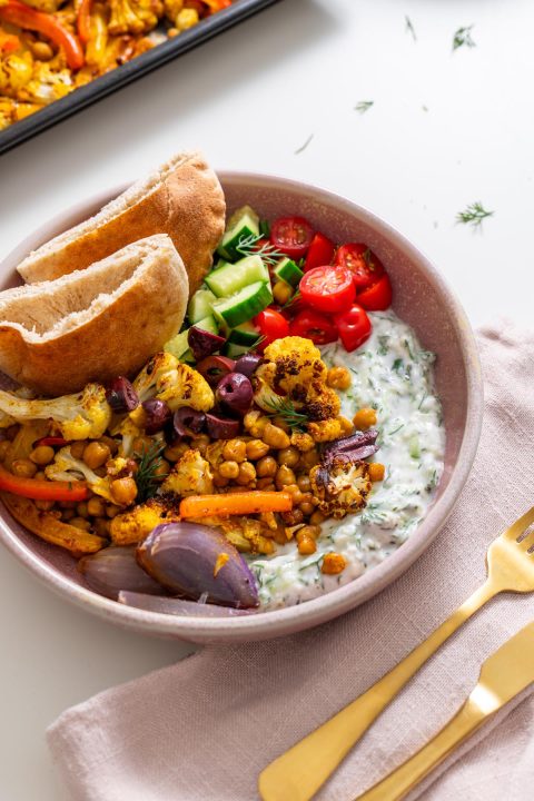 Bowl of Mediterranean sheet pan pita bowls with tzatziki, roasted veggies, olives, and pita on a light pink napkin.