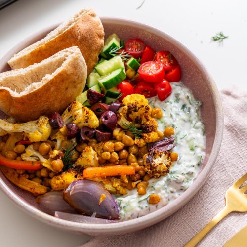 Bowl of Mediterranean sheet pan pita bowls with tzatziki, roasted veggies, olives, and pita on a light pink napkin.