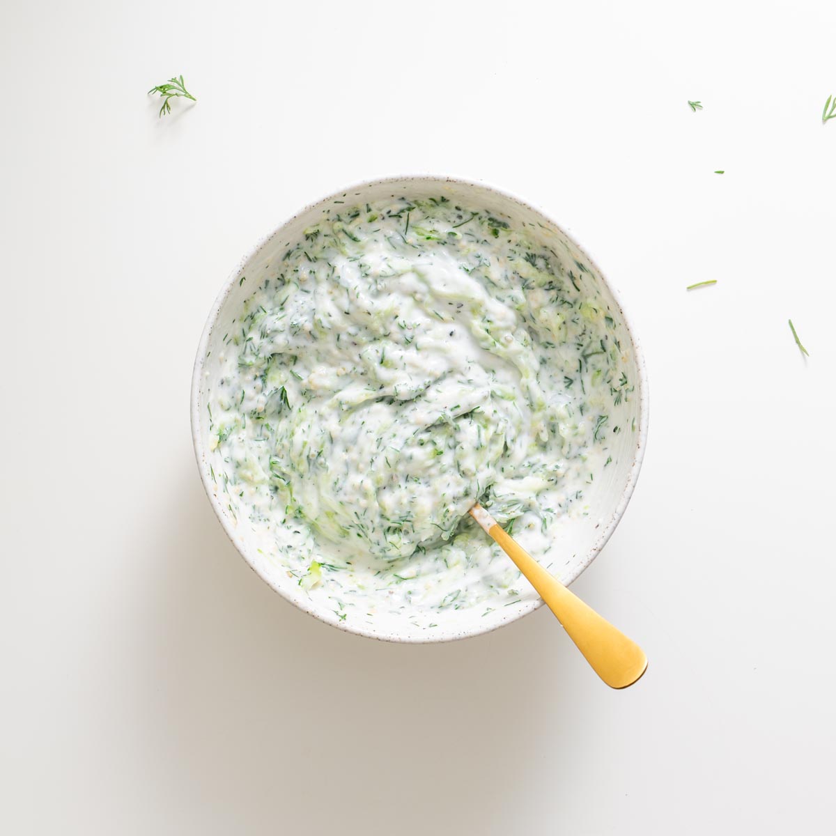 Creamy dairy-free tzatziki in a bowl with a gold spoon on a white surface.