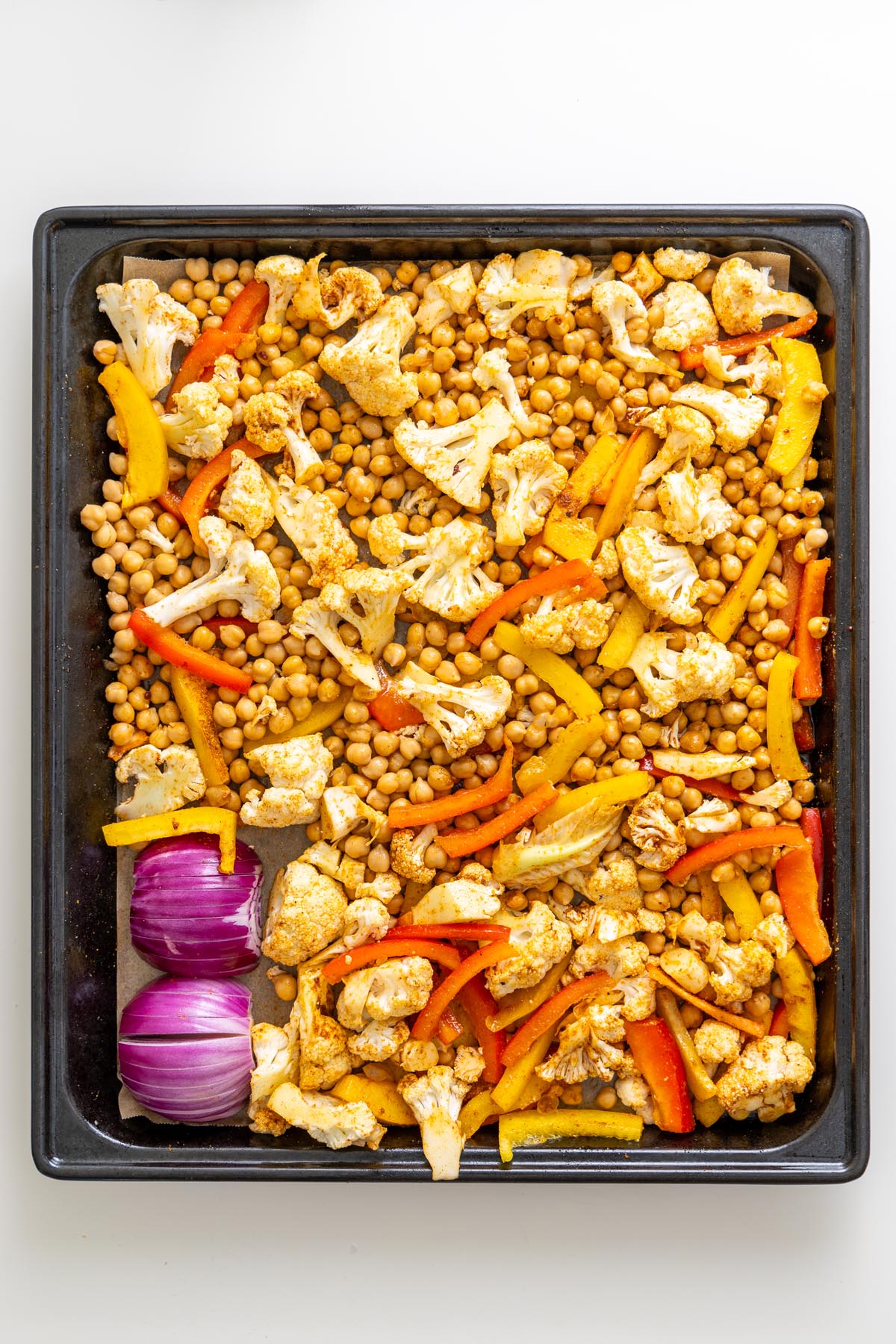 Unbaked chickpeas, cauliflower, red onion, and bell peppers on a sheet pan before roasting.