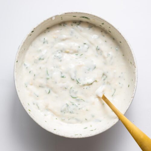 Creamy garlic yogurt sauce mixed with parsley in a white bowl with a gold spoon.