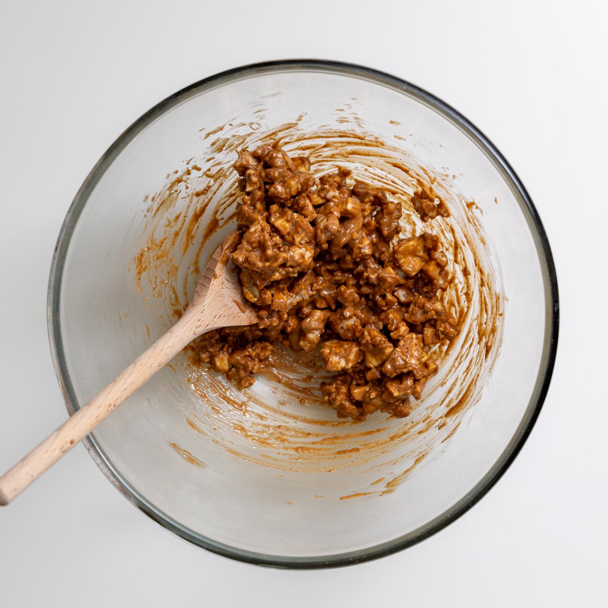 Glass bowl containing mixture of crumbled rice cakes, dried apple slices, almond butter and cinnamon with a wooden spatula