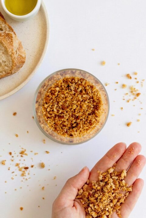 A bowl of golden homemade sourdough breadcrumbs with Sarah Cobacho holding some crumbs above a white surface