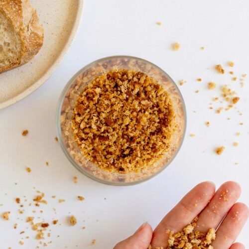 A bowl of golden homemade sourdough breadcrumbs with Sarah Cobacho holding some crumbs above a white surface
