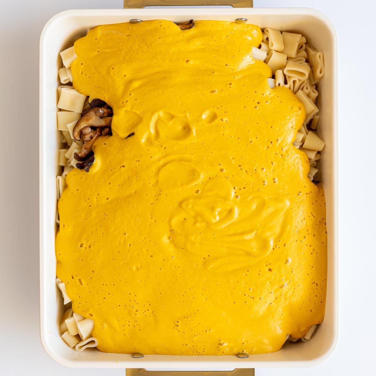 Bright orange lentil cheese sauce poured over pasta and mushrooms in a baking dish.