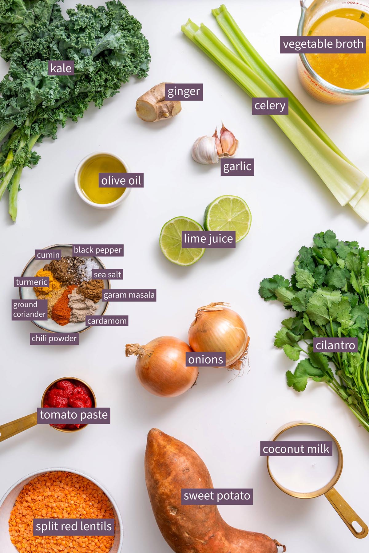 Vibrant ingredients for creamy red lentil and sweet potato soup on a white surface, including kale, ginger, garlic, celery, onions, spices, and lime.
