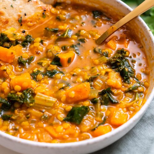Close-up of creamy red lentil and sweet potato soup with kale in a speckled bowl and gold spoon.