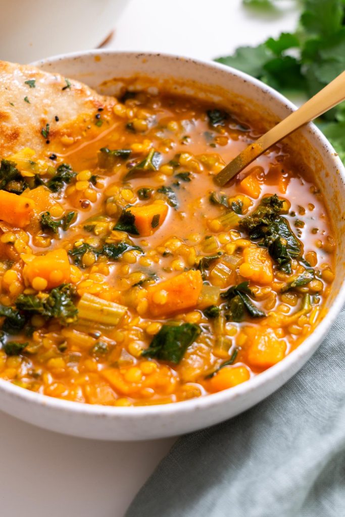 Close-up of creamy red lentil and sweet potato soup with kale in a speckled bowl and gold spoon.